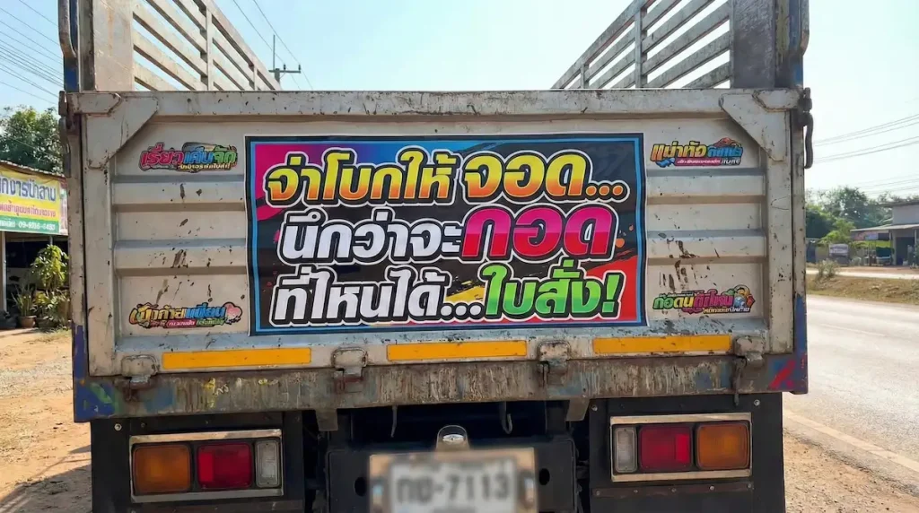 ภาพท้ายรถสิบล้อติดสติกเกอร์คำคมฮาๆ ว่า "จ่าโบกให้จอด นึกว่าจะกอด ที่ไหนได้ ใบสั่ง" (เบลอป้ายทะเบียนแล้ว) ประกอบบทความแคปชั่นคนขับรถสู้ชีวิต
